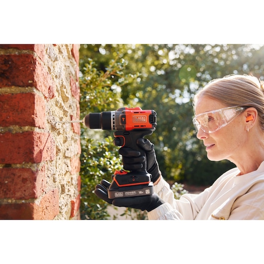 BLACKDECKER BCD383D1 cordless drill in use on a brick wall, outdoors, held by a person wearing gloves.