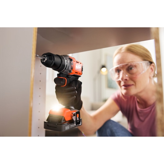 Person using a BLACK+DECKER cordless drill on furniture, wearing safety glasses and gloves.