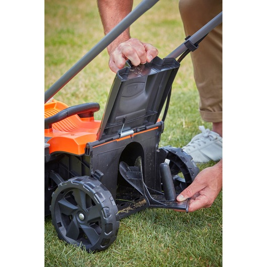 Photo of BLACKDECKER BCMW33184L2 lawn mower rear compartment open, showing wheel and user adjusting cover on grass.