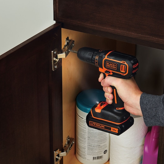 BLACKDECKER BDCDC18KB cordless drill used to fix cabinet hinge indoors, orange and black design.