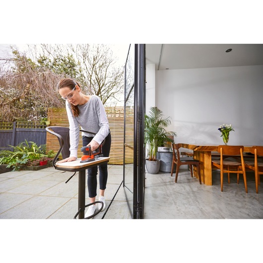 BLACKDECKER BDCDS18 sander on tabletop outdoors, person sanding wood, modern patio and dining area visible.