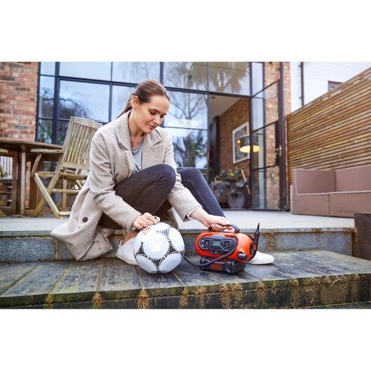 A person sitting outside on steps uses a BLACK+DECKER BDCINF18N air inflator to inflate a soccer ball. The portable orange and black inflator is placed next to the ball.