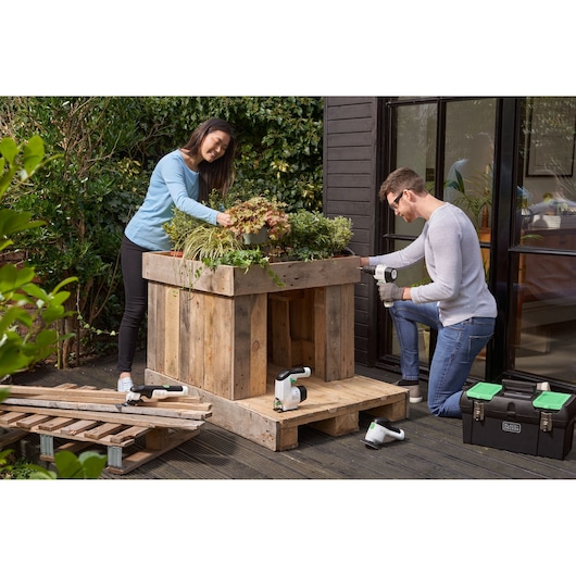 BLACKDECKER BDST60120 toolbox on patio with two people building a wooden planter, assorted power tools visible.