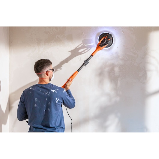Person sanding a wall with an orange BLACK+DECKER power sander. Wearing mask and safety glasses.