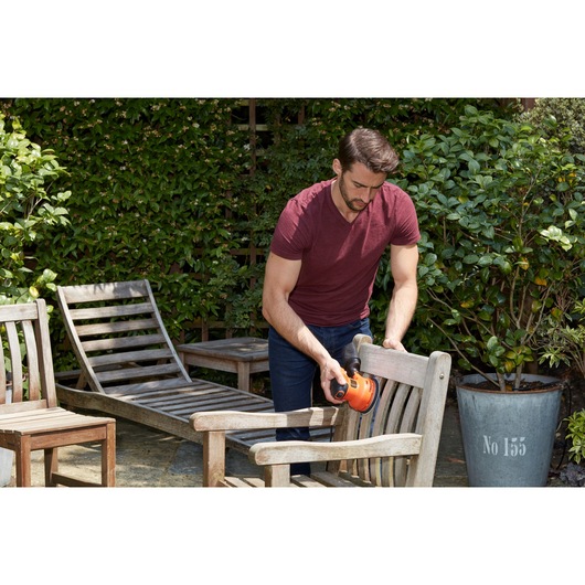 Person using BLACKDECKER BEW210 sander on wooden outdoor chair in garden setting.