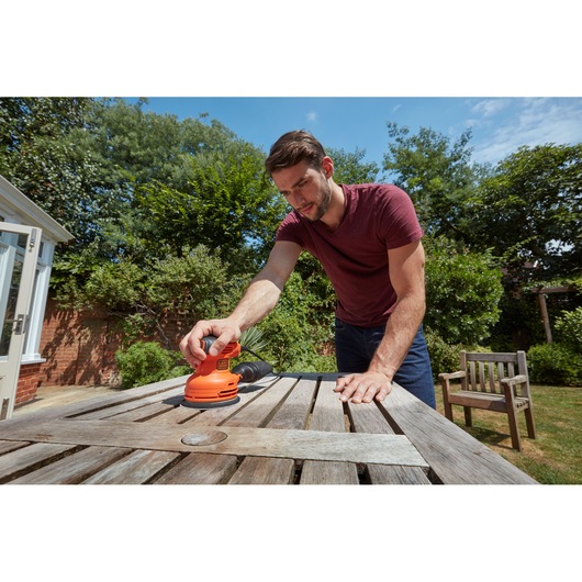 BLACK+DECKER BEW210 random orbital sander used to sand a weathered outdoor wooden table in a garden.