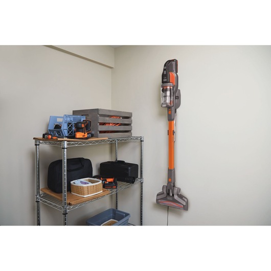 BLACKDECKER BHFEV182C cordless stick vacuum, orange and gray, wall-mounted next to metal shelving with tools.