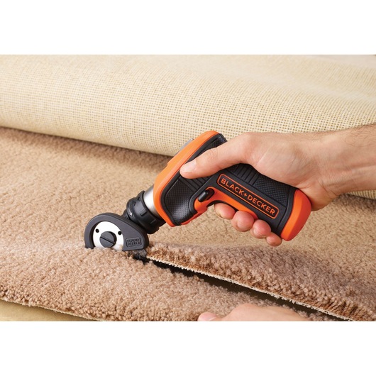 A person is using a BLACK+DECKER electric cutting tool to cut through a piece of beige carpet. The tool has a black and orange handle and is designed for precise cutting.