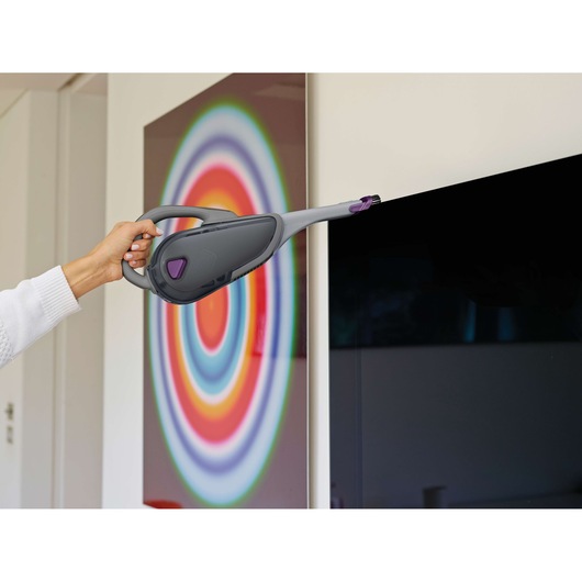 BLACKDECKER DVJ325BFS handheld vacuum with long nozzle cleaning TV screen, modern interior background.