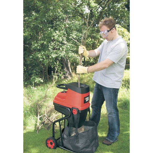 Photo of BLACK+DECKER GS2400 garden shredder in use outdoors, person feeding branches into machine, grass and trees visible.