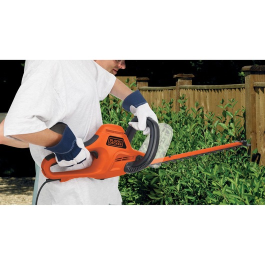 Photo of BLACK+DECKER GT6030 hedge trimmer in use by a person wearing gloves, trimming green bushes near a wooden fence.