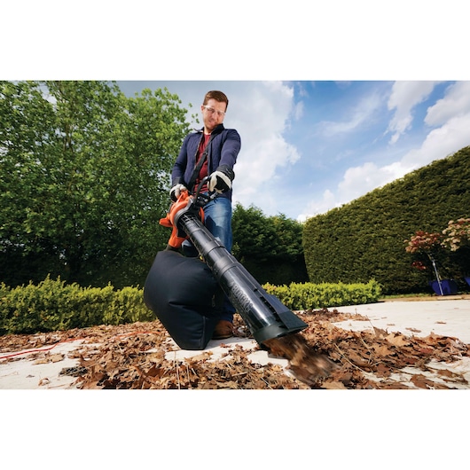 A person using a BLACK+DECKER leaf blower and vacuum to clear dry leaves from a patio area, with trees and hedges in the background.