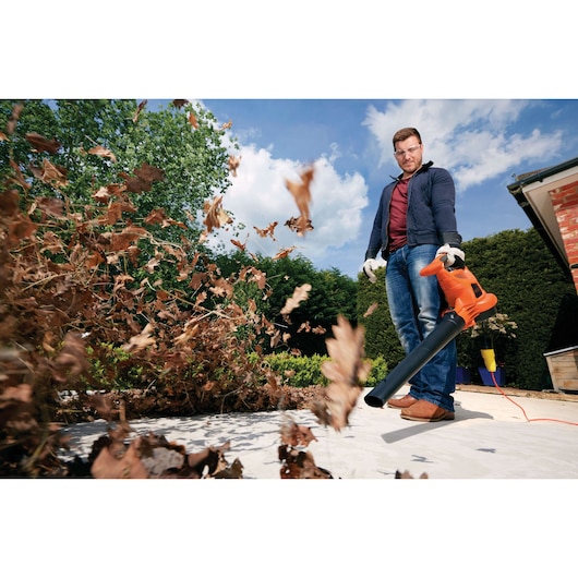 A person using an orange BLACK+DECKER leaf blower outdoors to clear fallen leaves, with leaves being blown away on a paved area near hedges and a brick building. The person’s face is intentionally blurred.
