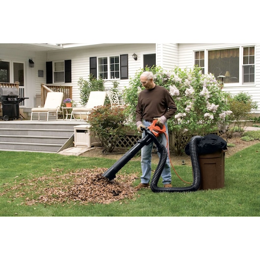 A person using a BLACK+DECKER garden vacuum and leaf blower to collect fallen leaves in a residential yard. The machine has a hose connected to a trash bin for collecting the leaves. The background shows a white house with a porch, outdoor furniture, and garden plants.