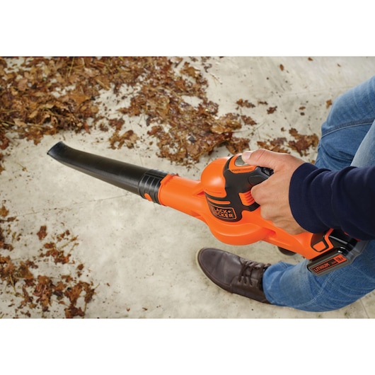A person is using an orange and black BLACK+DECKER cordless leaf blower to clear dry leaves from a concrete surface.
