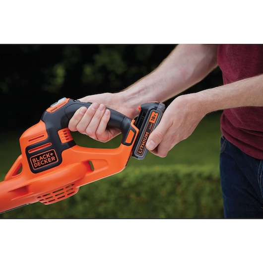 Close-up of a person inserting an 18V lithium battery into a BLACK+DECKER orange and black cordless power tool outdoors.