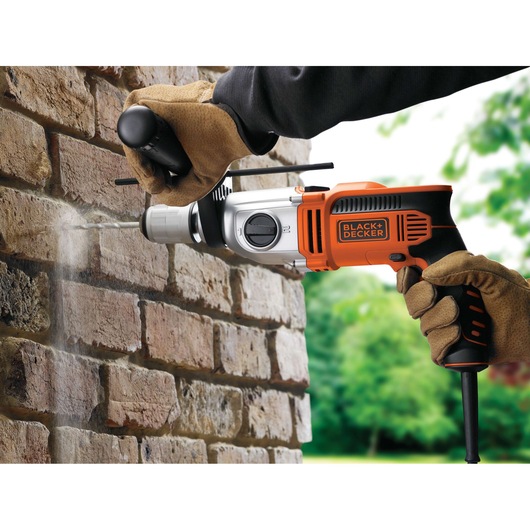 BLACK+DECKER KR911K hammer drill in use on a brick wall, orange and black design, user wearing gloves.