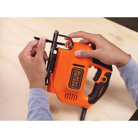BLACK+DECKER KS701E jigsaw held by two hands on light wood surface, showing close-up of blade attachment.