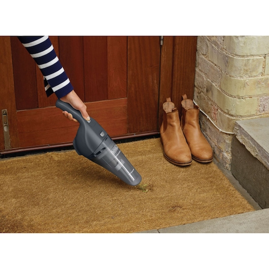 BLACK+DECKER NVB215WA handheld vacuum cleaner being used on a doormat near tan boots by a wooden door.