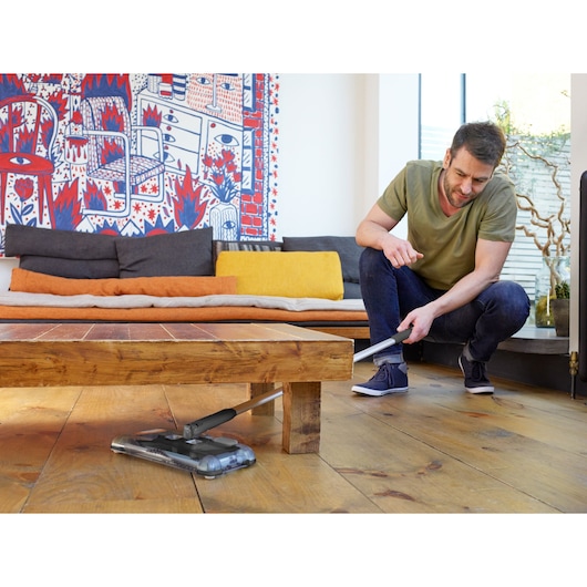 BLACKDECKER PSA215B cordless floor sweeper cleaning under wooden coffee table in modern living room.