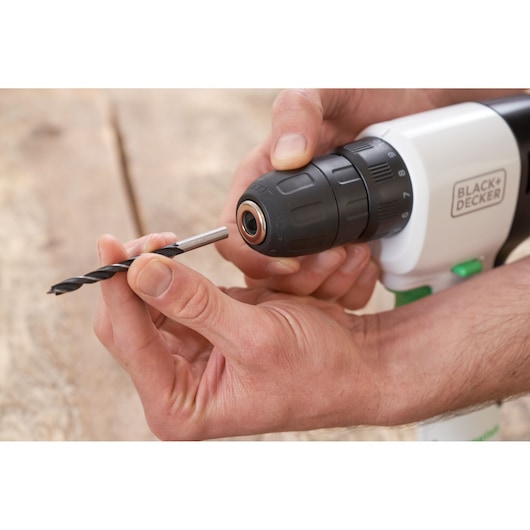 Hands inserting drill bit into BLACK+DECKER REVDD12C cordless drill, close-up view on wooden surface.