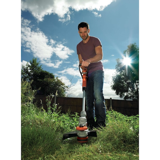 A person using a BLACK+DECKER STC1820 grass trimmer outdoors in a garden, wearing a purple t-shirt and jeans. The trimmer is cutting tall grass, with a wooden fence and trees in the background under a sunny sky.