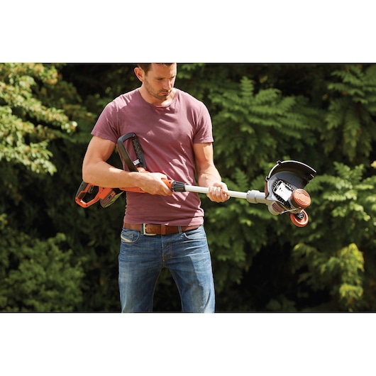 A person holding a BLACK+DECKER cordless grass trimmer outdoors, with greenery in the background. The person's face is blurred for privacy.