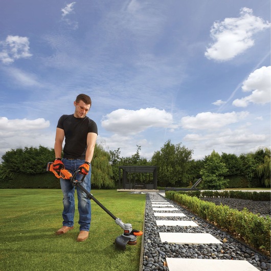 A person wearing protective gloves uses a BLACK+DECKER cordless grass trimmer (model STC1840) to trim the edge of a lawn near a garden path with stepping stones. The scene is outdoors with vegetation and a blue sky with scattered clouds.