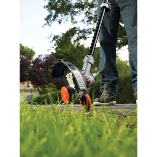 A person using a BLACK+DECKER cordless grass trimmer to edge along a lawn. The tool features orange and black wheels and is being used near a paved pathway, with green grass and trees in the background.